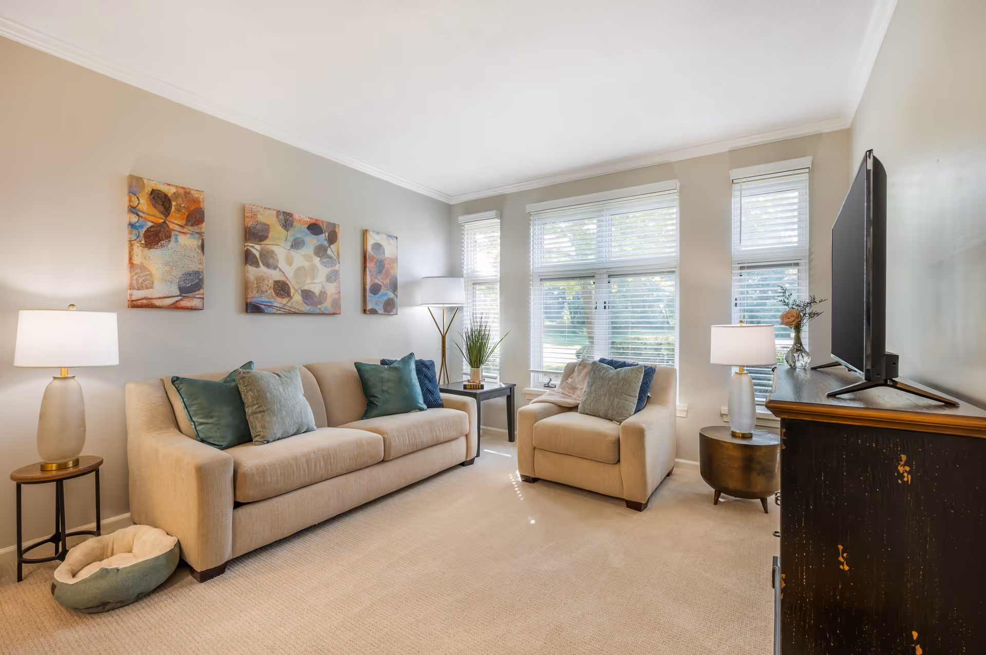 A cozy living room with a beige sofa and matching armchair, both adorned with green and blue throw pillows. There are three abstract leaf-themed paintings on the wall above the sofa. The room is well-lit with natural light coming through three large windows with white blinds. Two table lamps and one floor lamp provide additional lighting. A small pet bed is placed on the floor next to the sofa. A flat-screen TV sits on a dark wooden cabinet with a small vase of flowers beside it.