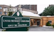 Entrance to Brooklyn United Methodist Church Home and Adult Day Healthcare facility with a green sign in front that reads 'Welcome to Brooklyn United Methodist Church Home and Adult Day Healthcare'. The building has a covered entryway and is surrounded by trees and other buildings.