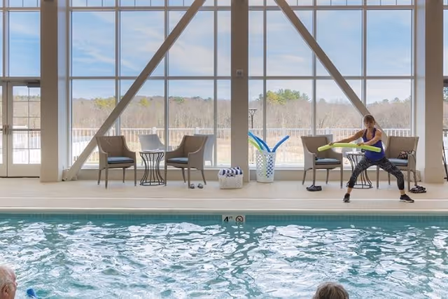 Indoor swimming pool with floor-to-ceiling windows, seating along the deck, and an instructor leading an exercise class using pool noodles.