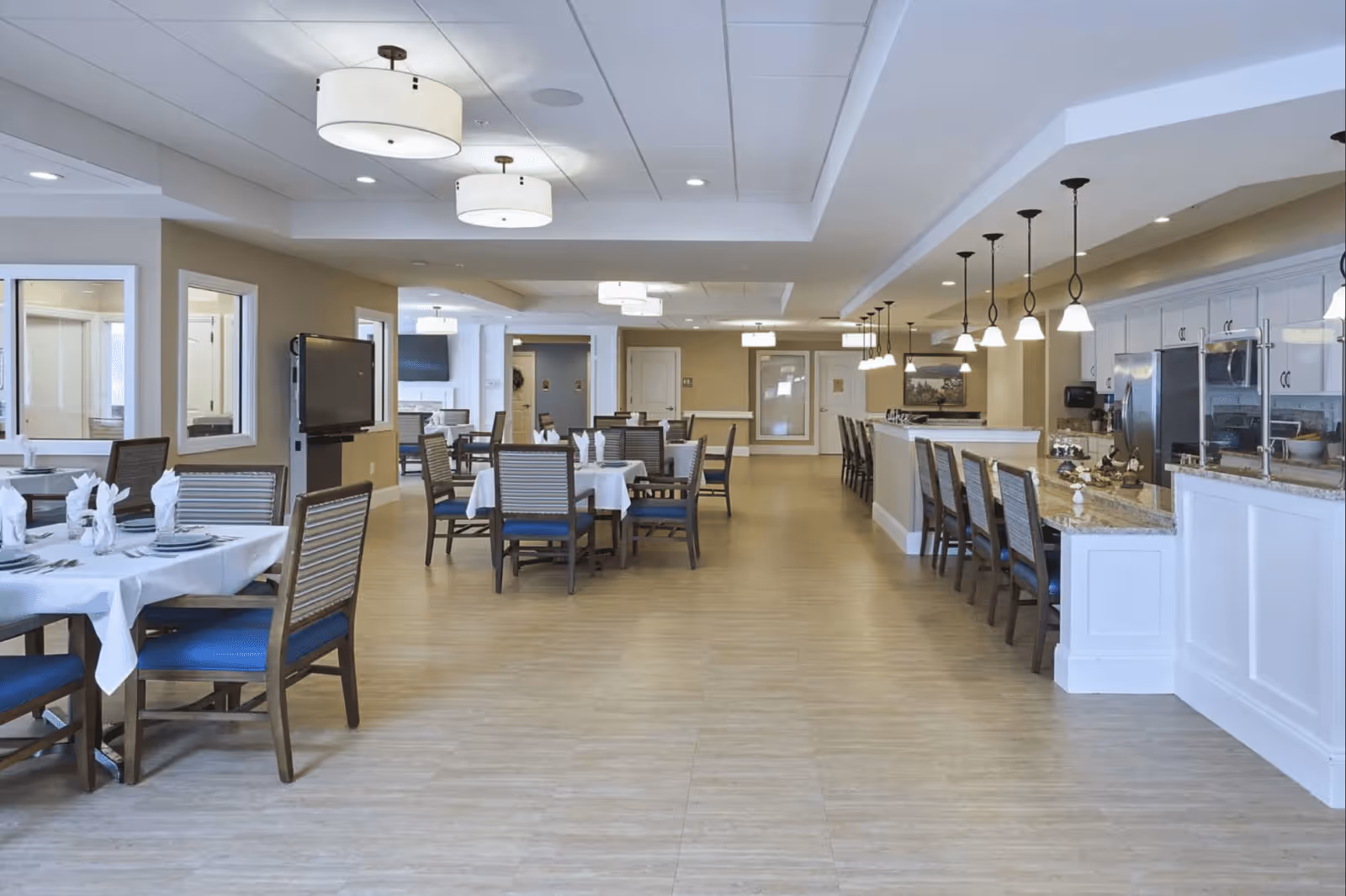 A spacious and well-lit dining area in a senior living facility featuring multiple tables with white tablecloths and neatly arranged chairs with blue cushions. The room has modern ceiling lights, a flat-screen TV mounted on the wall, and a long counter with bar stools and pendant lights above it. The flooring is light wood, and the walls are painted in neutral tones.