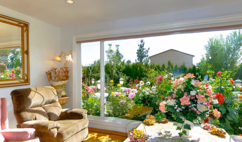 A cozy living room area with a comfortable brown armchair and a pink chair partially visible. There is a large window showing a vibrant garden with various colorful flowers and greenery outside. A table near the window holds a large floral arrangement and some small decorative items. The room is well-lit with natural light and has a wall-mounted lamp and a framed mirror on the wall.