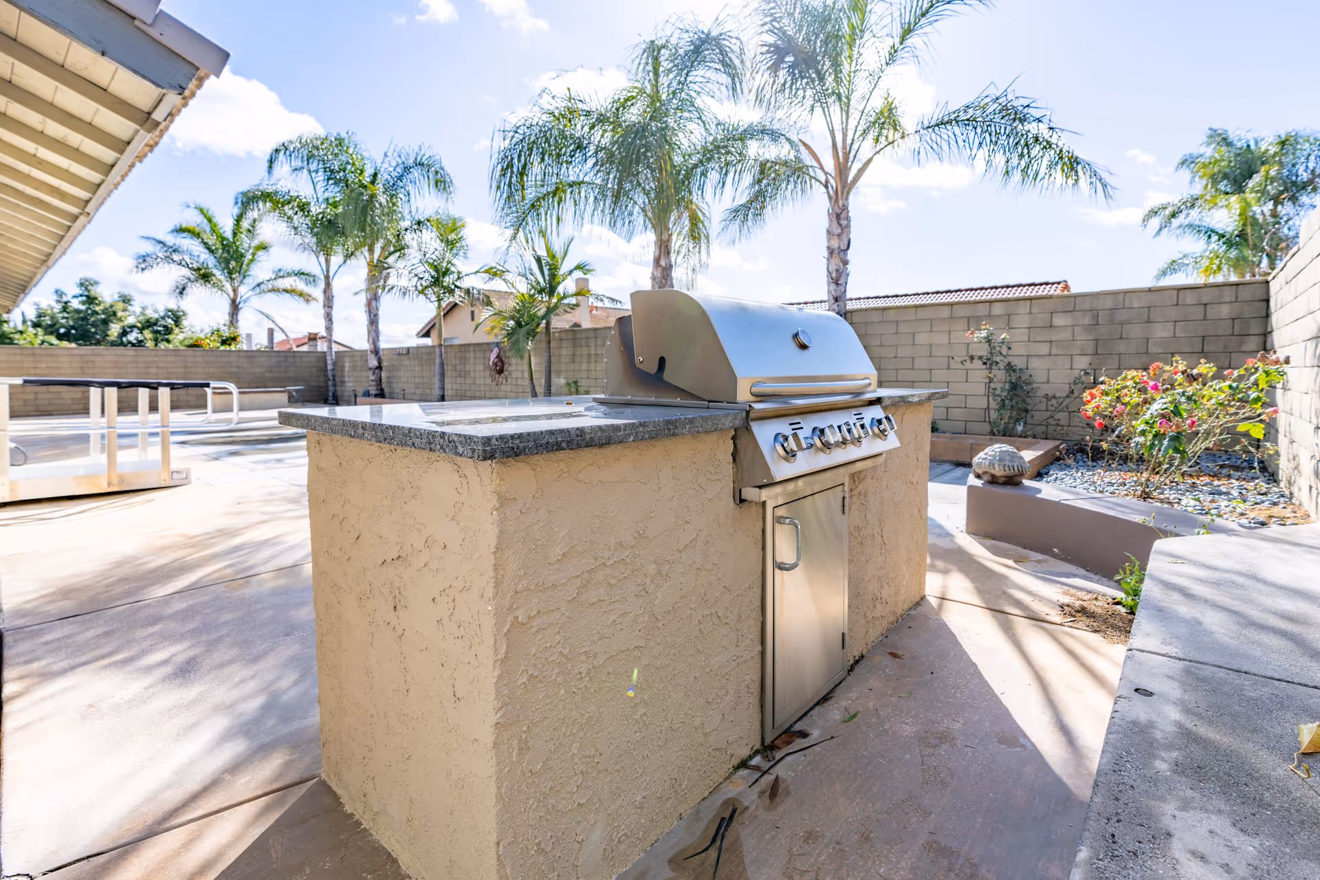 Outdoor patio area with a built-in stainless steel barbecue grill, surrounded by palm trees and a concrete block wall. There are some plants and flowers in a garden bed to the right, and a clear blue sky with some clouds overhead.