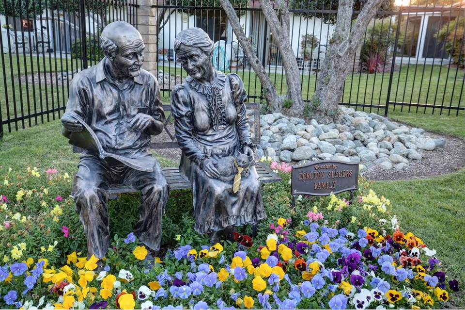 Bronze statue of an elderly man and woman sitting on a bench surrounded by colorful flowers. The man is holding a newspaper, and the woman has a cat with a yellow tie on her lap. Behind them is a black metal fence and a tree with rocks around its base. A small plaque reads 'Donated by Dorothy Slegers Family.'