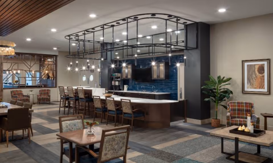 Modern communal dining and lounge area with a long bar island, pendant lights, tables and chairs, and lounge seating.