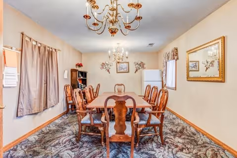 A dining room with a long wooden table surrounded by eight matching wooden chairs with cushioned seats. The room has beige walls, two chandeliers hanging from the ceiling, a large mirror on the right wall, a window with curtains, and a white refrigerator in the back corner. There is a patterned carpet on the floor and decorative wall art above the refrigerator.