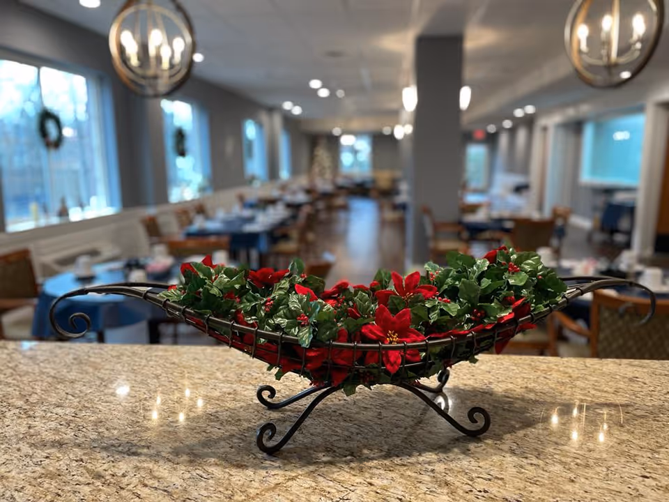 A decorative centerpiece with red poinsettias and green leaves in a black metal holder placed on a granite countertop. In the background, a dining area with tables and chairs is visible, softly lit with hanging lights and natural light from windows.