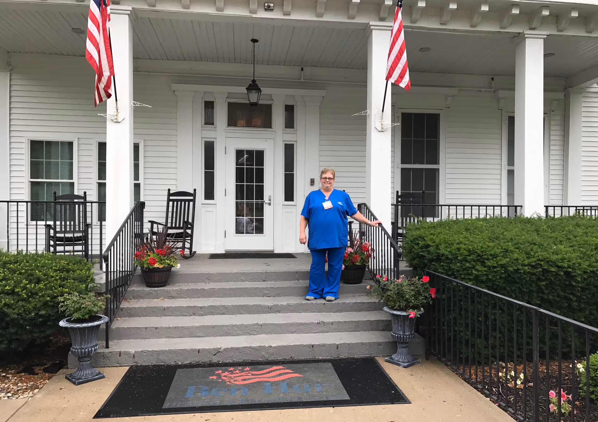 A healthcare worker in blue scrubs stands on the steps of a white building entrance with two American flags mounted on the porch columns. There are black rocking chairs and flower pots on either side of the steps, and a welcome mat with a red and blue design at the bottom of the stairs.