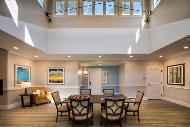 A bright and spacious common area with high ceilings and large windows near the ceiling letting in natural light. The room features a table with six chairs arranged around it, a comfortable armchair with a side table and lamp, and framed artwork on the walls. The walls are painted in neutral tones with a light blue accent wall near two white doors.