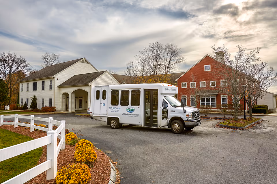 A white shuttle bus parked in front of a senior living facility with two buildings, one white and one red, surrounded by trees and landscaping under a cloudy sky.