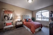 A cozy bedroom with a single bed dressed in a rust-colored comforter and pillows. The room features a nightstand with a lamp, a cushioned chair with a coral pillow, a large window with an arched top letting in natural light, a framed artwork on the wall, and a potted plant near the window.