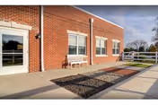 Exterior view of a brick building with three windows and a white door. A white bench is placed against the wall between the windows. The area in front of the building has a concrete walkway and a rectangular section filled with dark gravel. There is a white fence and some grass visible in the background under a partly cloudy sky.