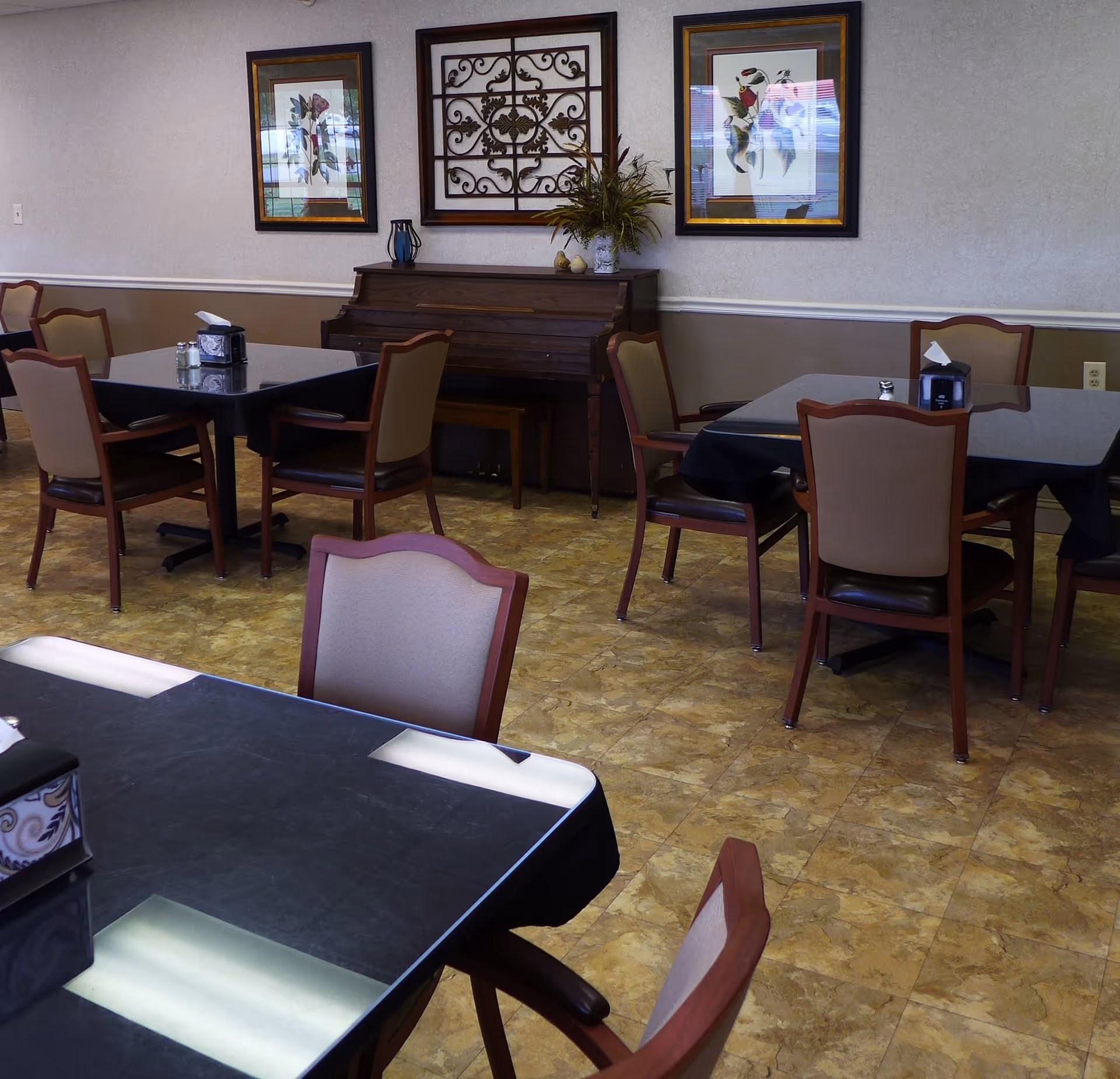 Interior view of a dining room with several tables and chairs arranged neatly. Each table has a black tablecloth and a tissue box. There is a piano against the wall decorated with a plant and ornamental wall art, along with framed pictures on the wall.