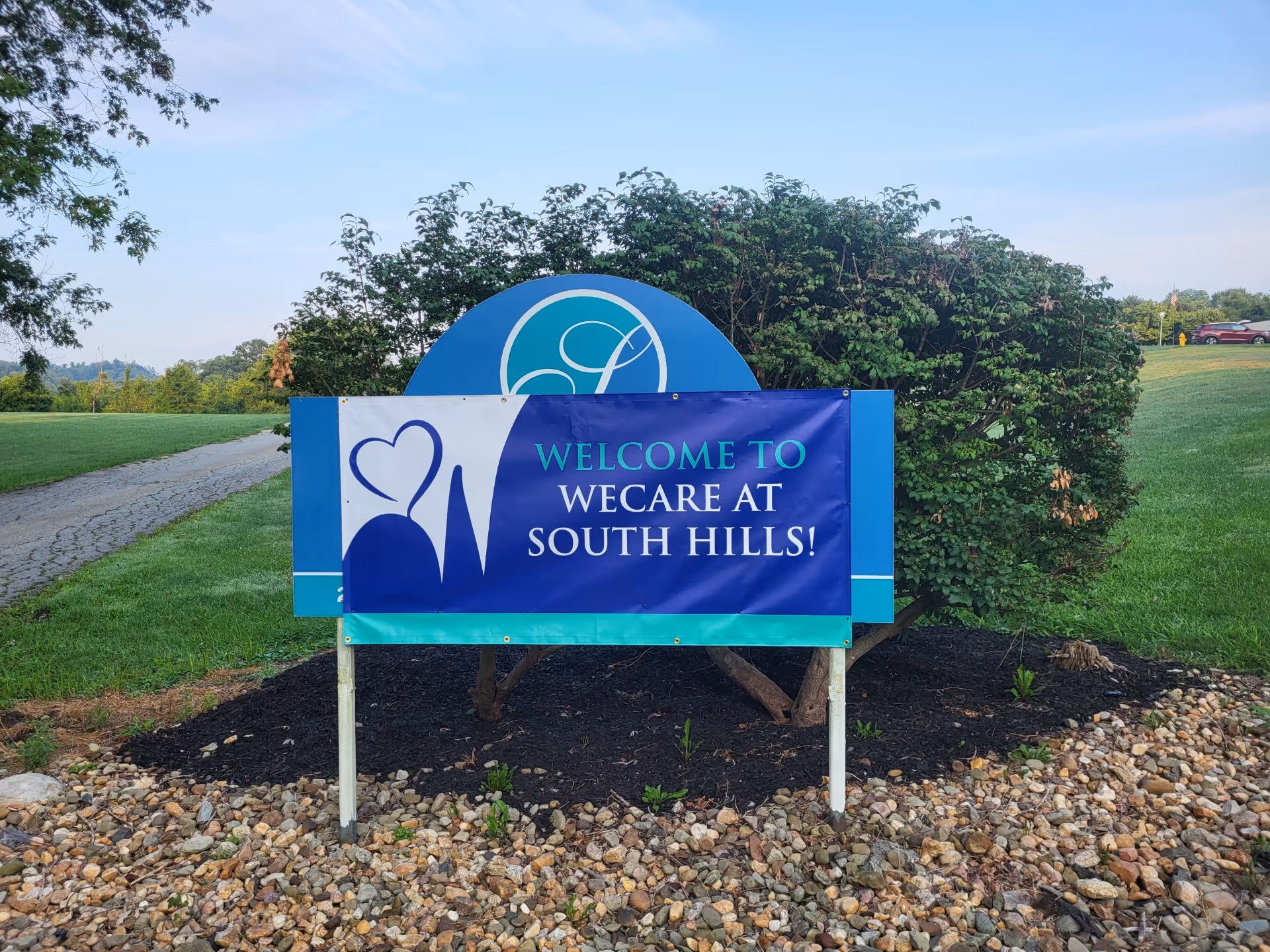 Outdoor sign with a blue and white banner that reads 'Welcome to WeCare at South Hills!' placed in a landscaped area with rocks and mulch, surrounded by green grass, trees, and a clear sky.