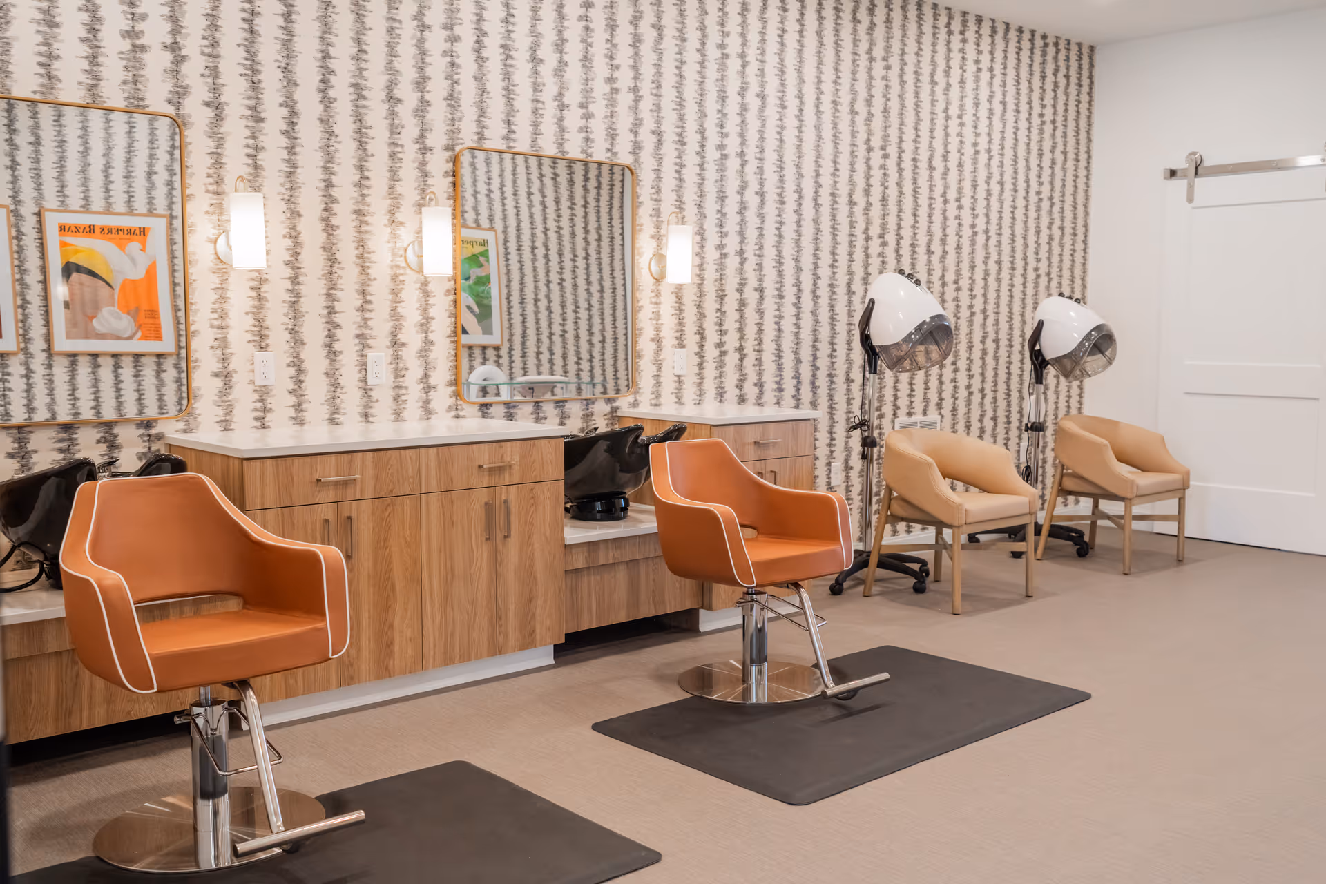 Interior view of a salon area in an assisted living facility featuring two orange salon chairs in front of mirrors mounted on a patterned wall, wooden cabinetry with sinks, and two beige chairs with hair dryers nearby.