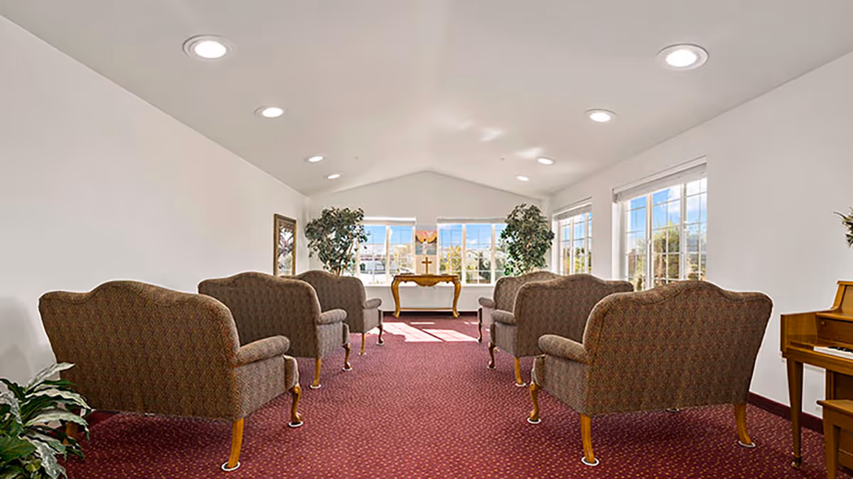 A bright room with six upholstered armchairs arranged in two rows facing a small wooden table with a cross on it. The room has large windows letting in natural light, potted plants on either side of the table, a piano on the right, and a red carpeted floor.