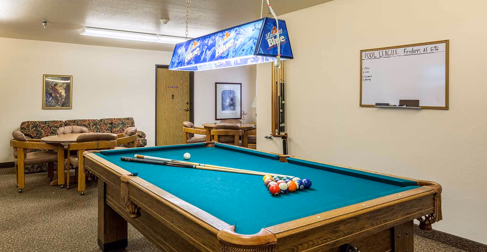 Indoor recreation room featuring a pool table with cues and racked balls, seating tables and a couch, and a wall-mounted whiteboard.