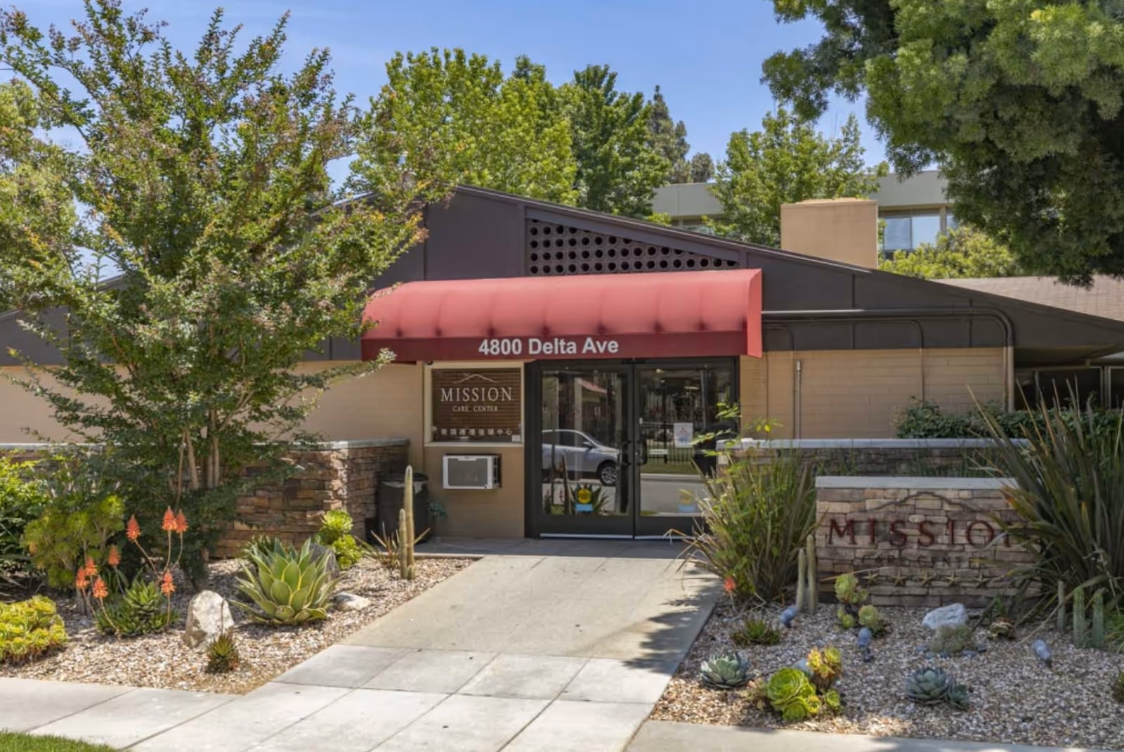 Entrance of Mission Care & Rehabilitation Center with a red awning displaying the address 4800 Delta Ave. The building is surrounded by landscaped plants and trees, with a stone sign partially visible on the right side.