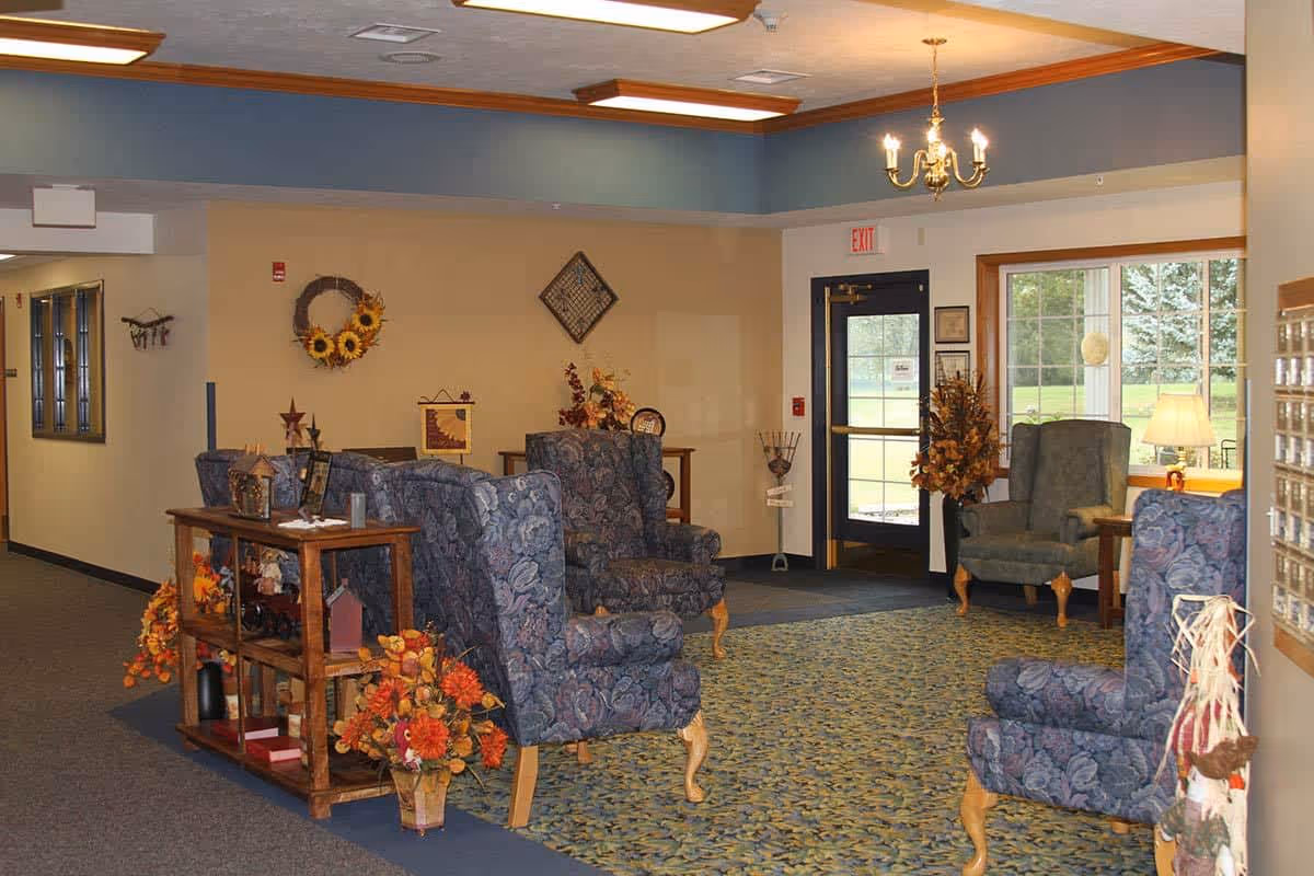 A cozy senior living facility common area with floral-patterned armchairs arranged around a carpeted floor. The room is decorated with autumn-themed floral arrangements and wall hangings, featuring a large window letting in natural light and a door marked with an exit sign.