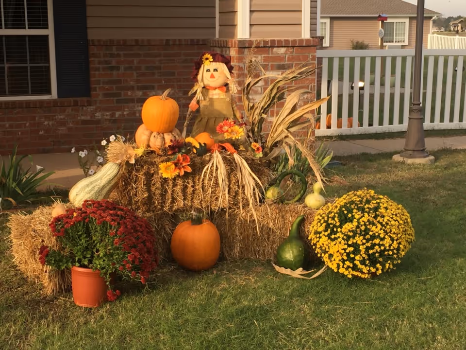 Autumn display of pumpkins, hay bales, gourds, potted mums and a scarecrow arranged on the front lawn of a brick house.