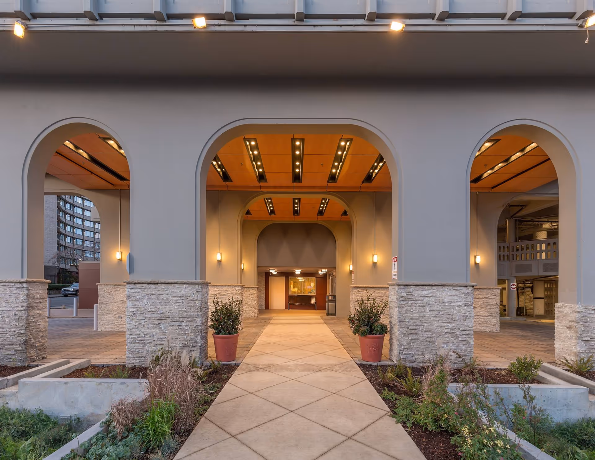 Entrance area of a building with three large archways, stone pillars, potted plants, and a tiled walkway leading inside. The ceiling inside the archways has wooden panels with recessed lighting.