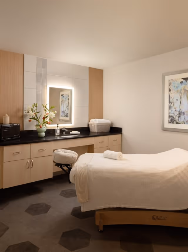 A serene treatment room with a massage table covered in a white blanket and a rolled towel on top. The room features a countertop with a sink, a vase with flowers, a basket with towels, and a backlit mirror above the sink. There is a framed abstract painting on the wall and a cushioned stool near the massage table.