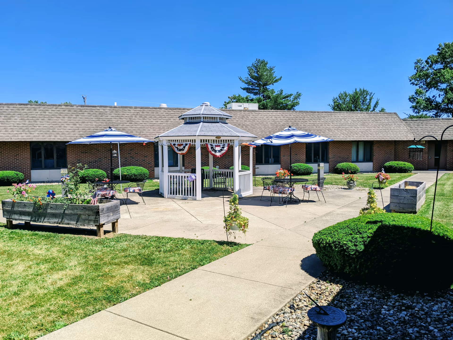 Outdoor courtyard area with a white gazebo decorated with red, white, and blue bunting. There are two blue and white striped umbrellas shading metal tables and chairs. Raised garden beds with flowers and greenery surround the paved area. The building in the background is a single-story brick structure with windows and a shingled roof. The sky is clear and blue.