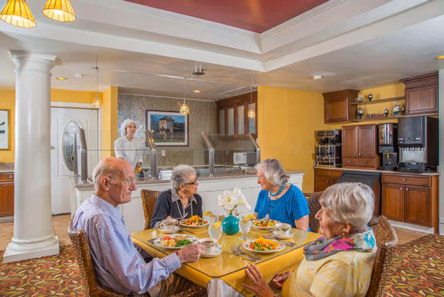Four elderly people sitting around a dining table in a well-lit assisted living facility dining room, enjoying a meal together. A staff member is visible behind a serving counter in the background. The room has warm yellow walls, wooden cabinetry, and decorative lighting.