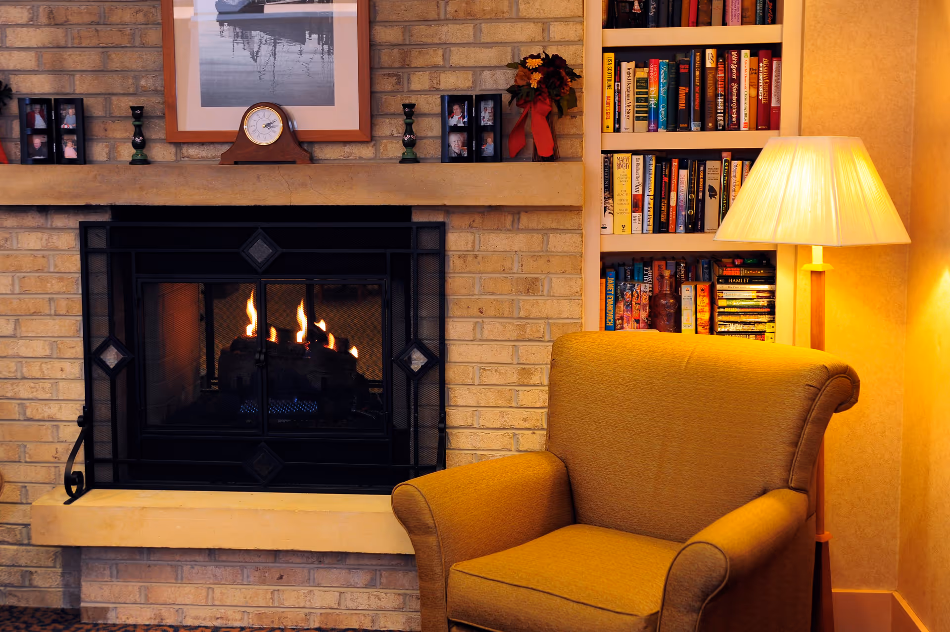 Cozy living room corner with a lit fireplace set in a brick wall, a mustard yellow armchair, a floor lamp with a beige shade, and a built-in bookshelf filled with books. The fireplace mantel holds a clock, framed photos, and decorative items.