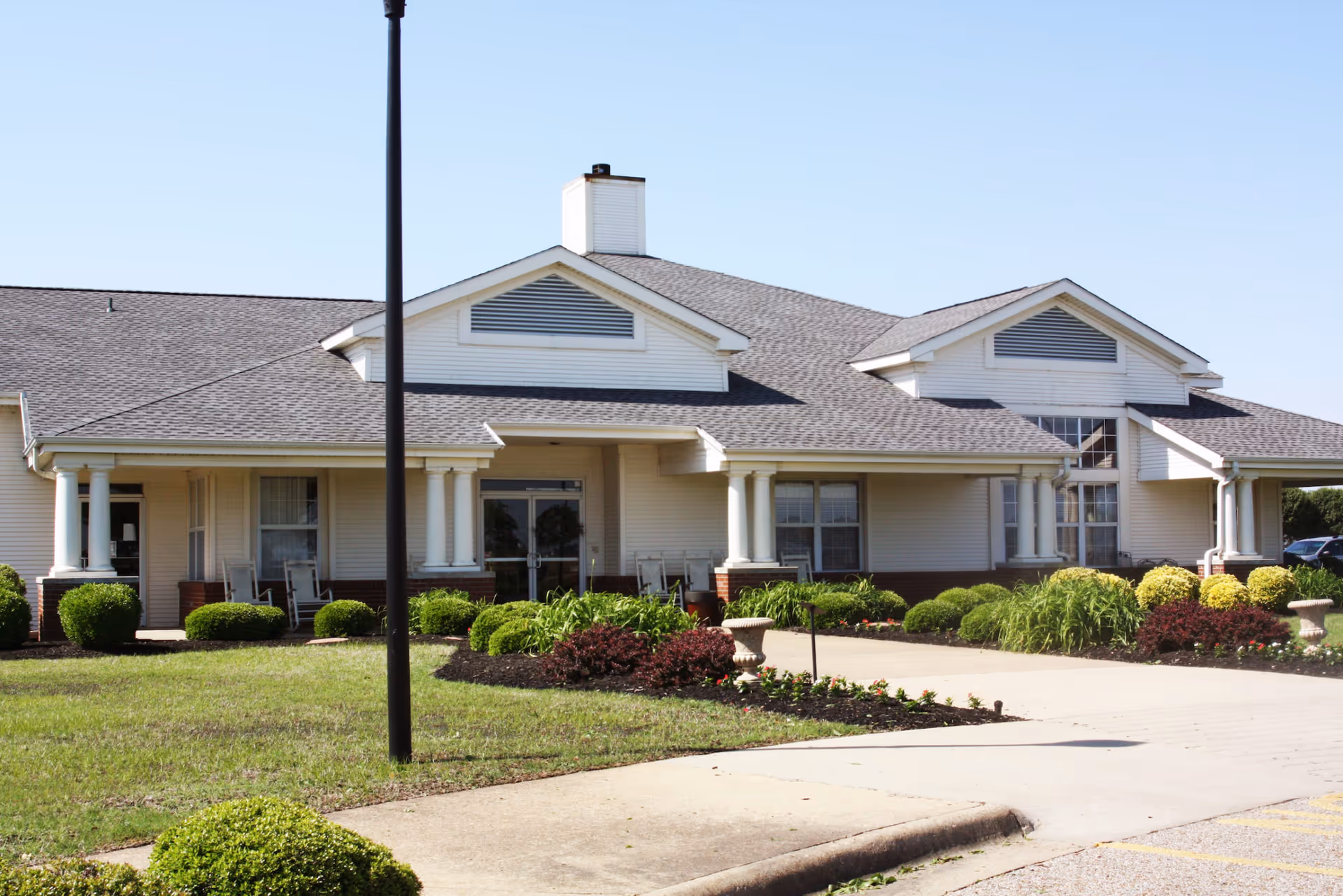 Exterior view of a single-story senior living facility building with a pitched roof, white siding, and brick accents. The entrance is covered by a small porch supported by white columns, with rocking chairs on either side. Well-maintained landscaping with green bushes and plants surrounds the walkway leading to the entrance. A clear blue sky is visible above.