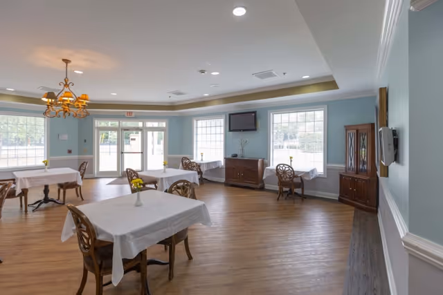 A bright dining room with wooden floors and light blue walls. Several tables covered with white tablecloths are arranged throughout the room, each with a small vase holding yellow flowers. Wooden chairs with intricate backs accompany the tables. Large windows allow natural light to fill the space. A chandelier hangs from the ceiling, and a flat-screen TV is mounted on the wall above a wooden cabinet.