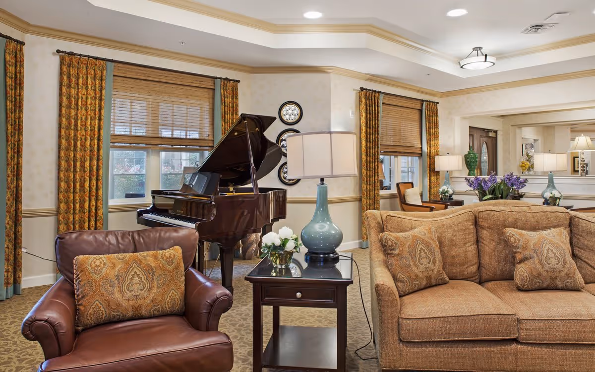 A cozy senior living room with a grand piano, upholstered sofa and armchair, side tables and lamps.