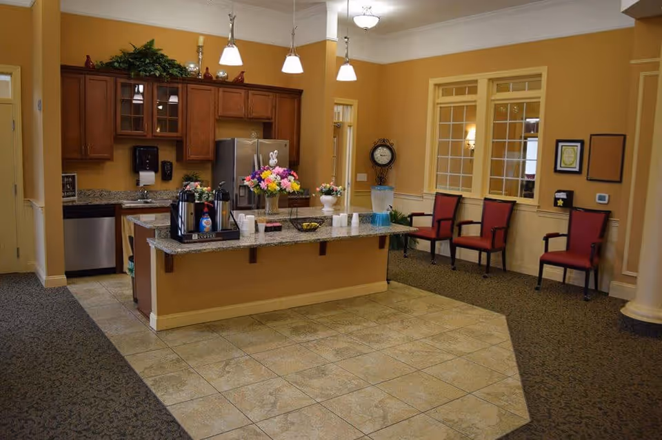 Interior view of a senior living facility kitchen area with a granite countertop island in the center. The island has coffee dispensers, cups, and flower arrangements on it. Behind the island are wooden cabinets, a stainless steel refrigerator, and a dishwasher. To the right, there are three red cushioned chairs against a wall with windows and framed decorations. The floor is a combination of tile and carpet, and the walls are painted a warm beige color.