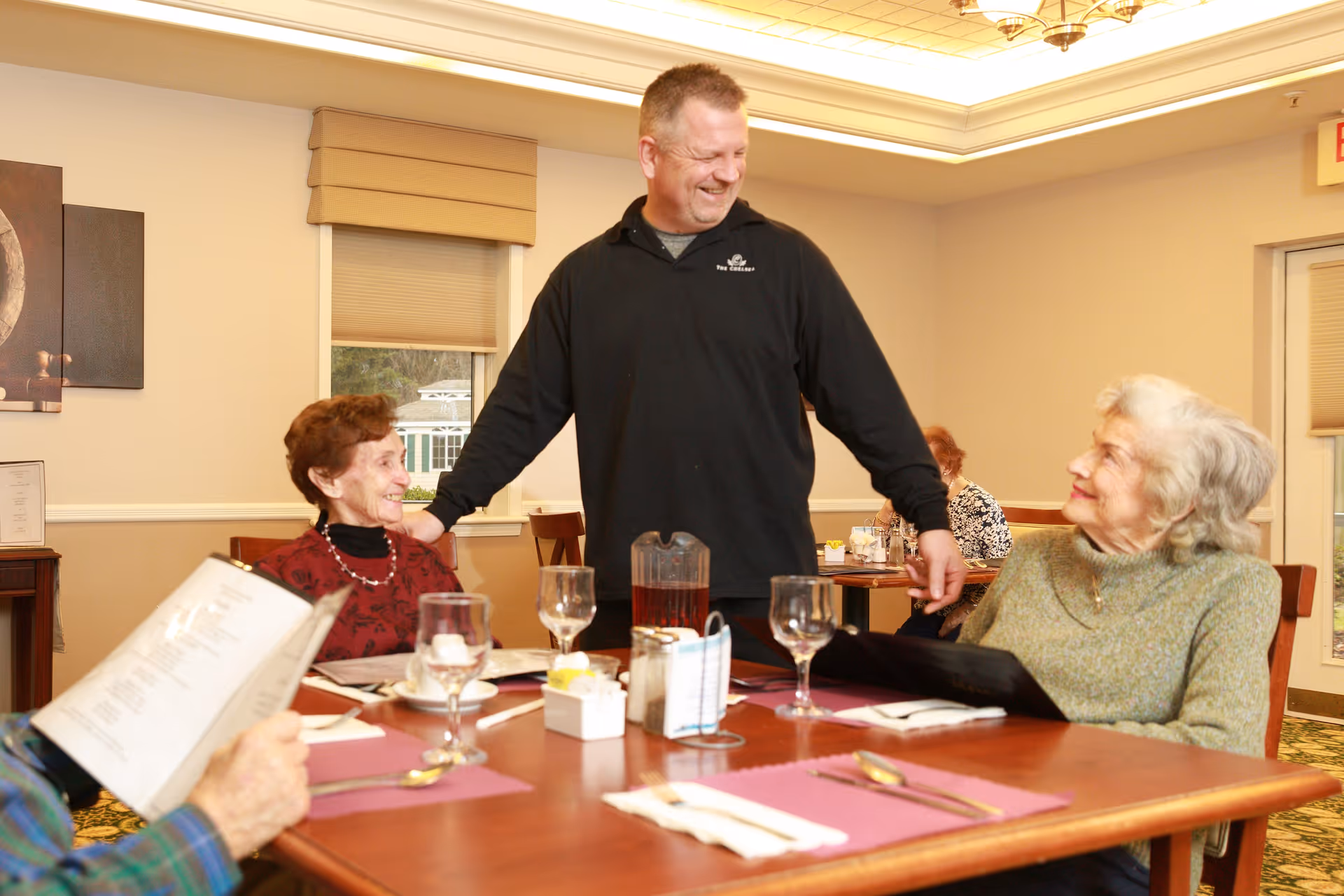 A man stands and smiles while interacting with two elderly women seated at a dining table in a well-lit room. The table is set with glasses, menus, and utensils. Another elderly person is partially visible holding a menu. The setting appears to be a communal dining area.