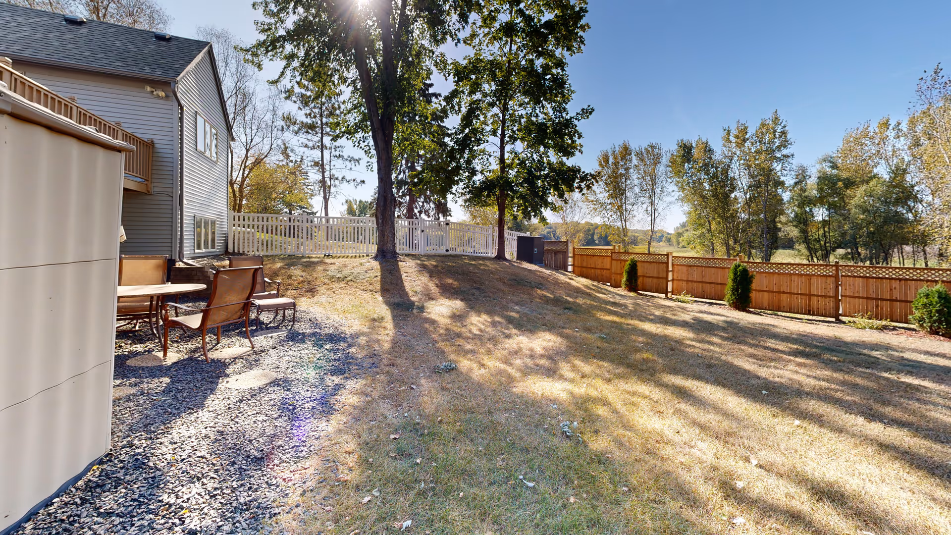 Sunlit backyard with patio furniture, a tree, and wooden fencing beside a two-story house.