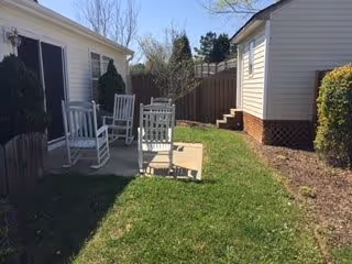 Small outdoor courtyard between two single-story buildings with a grass lawn, a concrete patio holding white rocking chairs, shrubs and a wooden fence.