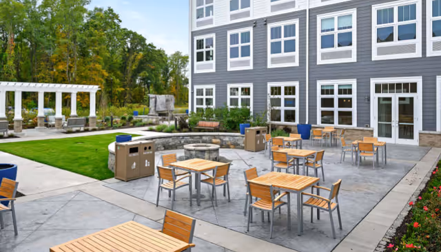 Outdoor patio area at a senior living facility with multiple wooden tables and chairs arranged on a concrete surface. There is a circular stone fire pit in the center, surrounded by benches. The building exterior is visible with multiple windows and doors. Green grass, a white pergola, and trees are in the background.