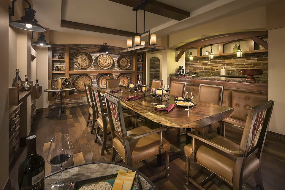 A cozy dining room with a long wooden table surrounded by leather chairs. The table is set with wine glasses, candles, and napkins. The room features warm lighting, brick accent walls, decorative plates on shelves, and a small bar area with pendant lights.
