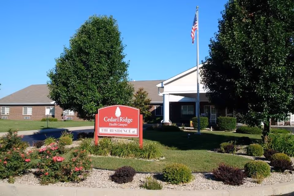 Exterior view of Cedar Ridge Health Campus showing a red sign with the facility name, surrounded by landscaped bushes and flowers. The building is visible in the background with a flagpole flying the American flag and trees on either side.