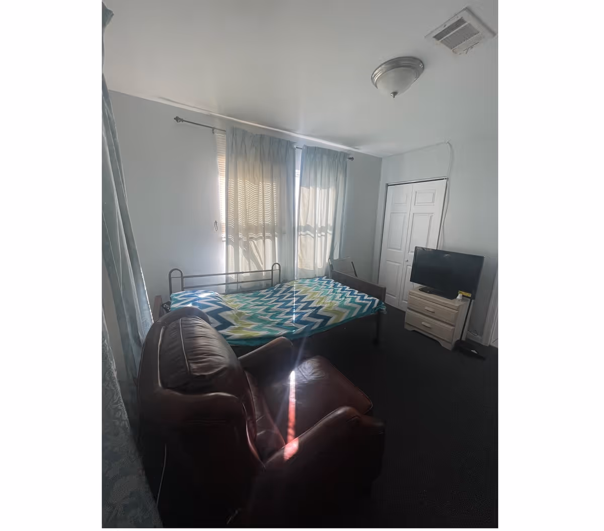 Small bedroom with a bed covered by a colorful chevron blanket, a brown leather armchair, a TV on a dresser, and curtained windows.