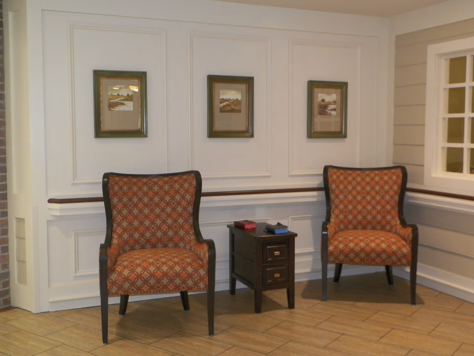 Two patterned armchairs with wooden frames are positioned on either side of a small dark wooden side table with two drawers. The chairs and table are set against a white paneled wall with three framed landscape paintings hanging above. The floor is tiled with light brown tiles, and there is a window with white framing on the right side.
