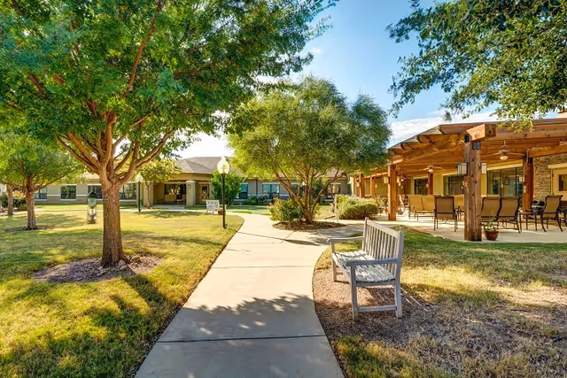 A sunny outdoor courtyard at The Auberge at Benbrook Lake featuring a paved walkway, green grass, several trees, a wooden bench, and a covered patio area with chairs and tables.