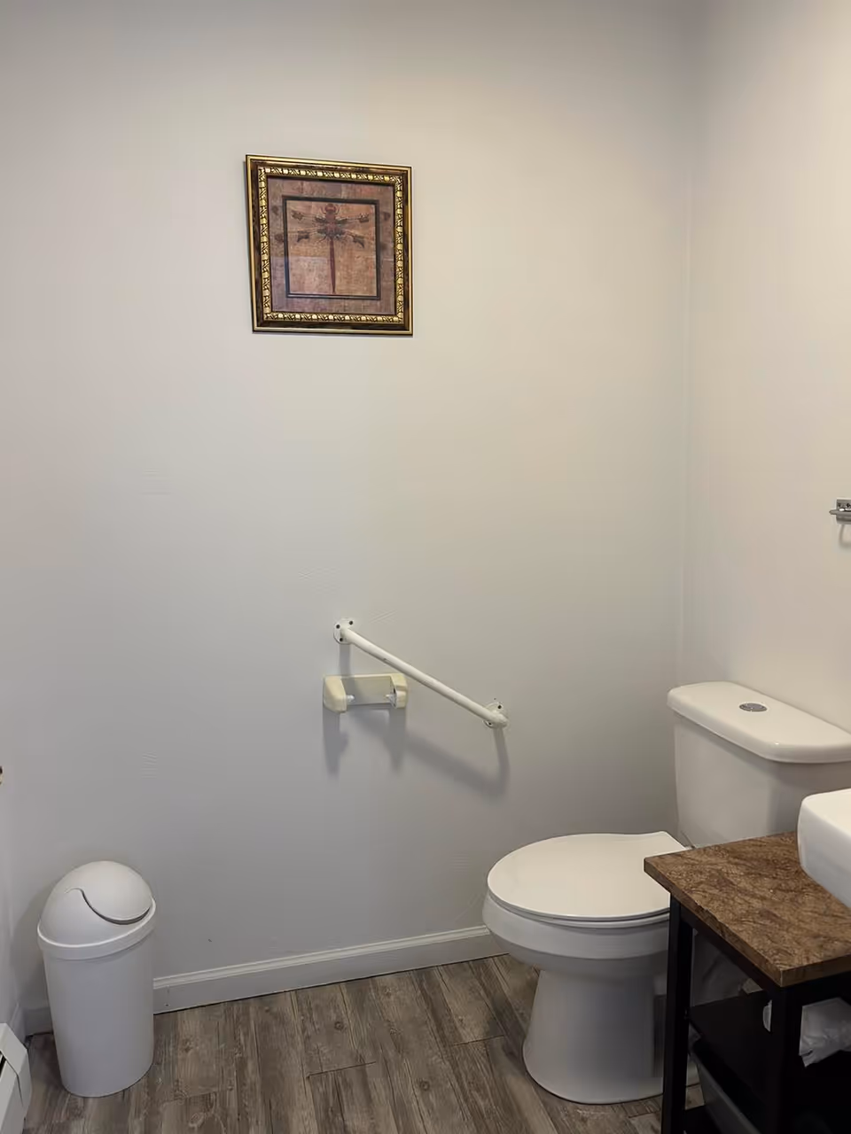 Small bathroom with a toilet, trash bin, grab bar, sink on a small vanity, and framed artwork on a white wall.
