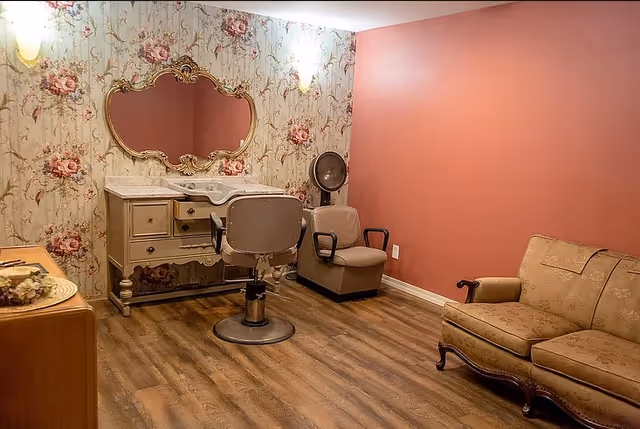 A cozy room with vintage-style furniture including a floral-patterned vanity with an ornate mirror, a salon chair, a hair dryer chair, and a beige upholstered loveseat. The room has floral wallpaper on one wall and a solid pink wall on the other, with wood flooring.