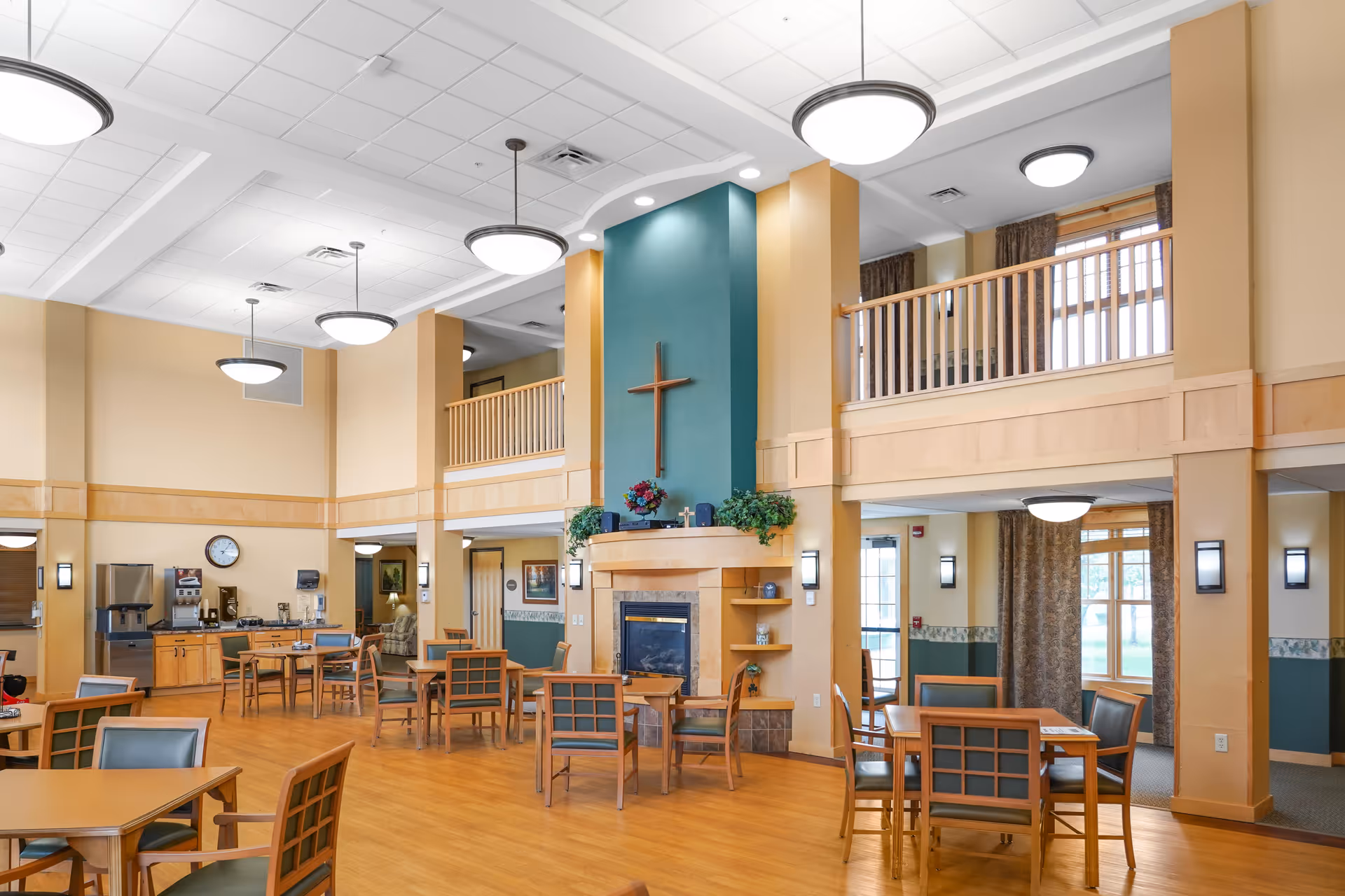 Spacious senior living common dining area with tables and chairs, a fireplace topped by a cross, high ceilings and an upper balcony.