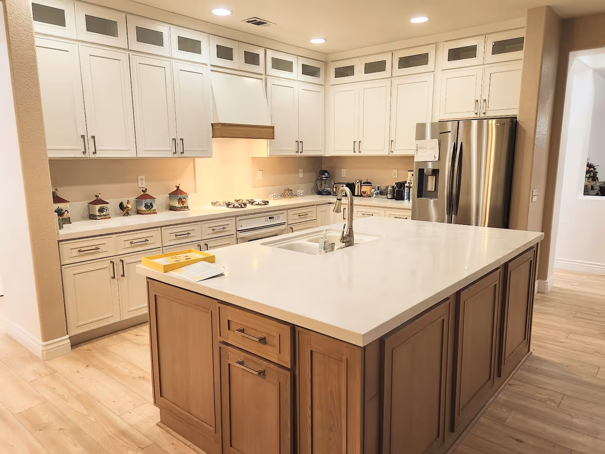 A spacious kitchen with white upper cabinets and light wood lower cabinets on the island. The kitchen features a large white countertop island with a built-in sink and faucet. Stainless steel refrigerator and built-in stove are visible, along with various kitchen appliances and decorative canisters on the counters. The floor is light wood, and the ceiling has recessed lighting.