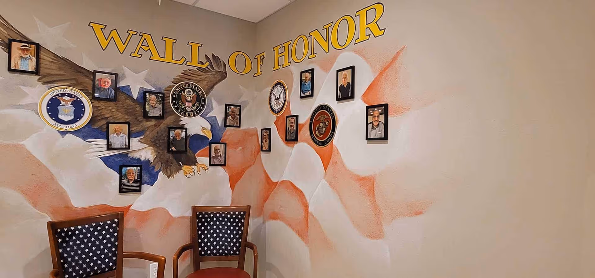 Interior wall featuring a 'Wall of Honor' mural with an eagle, American flag motif, military emblems and framed photos above two chairs.