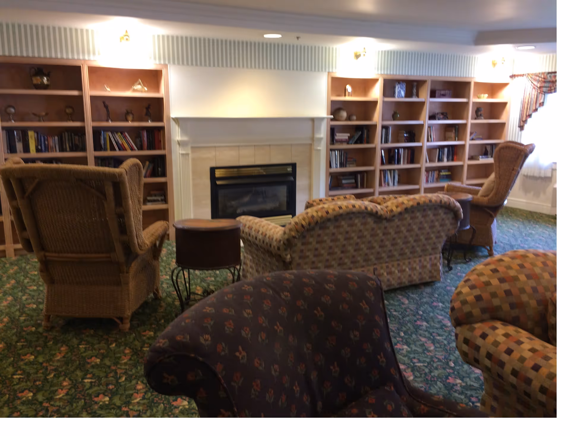 Cozy common room with a fireplace, built-in bookshelves and several upholstered and wicker chairs on a patterned carpet.