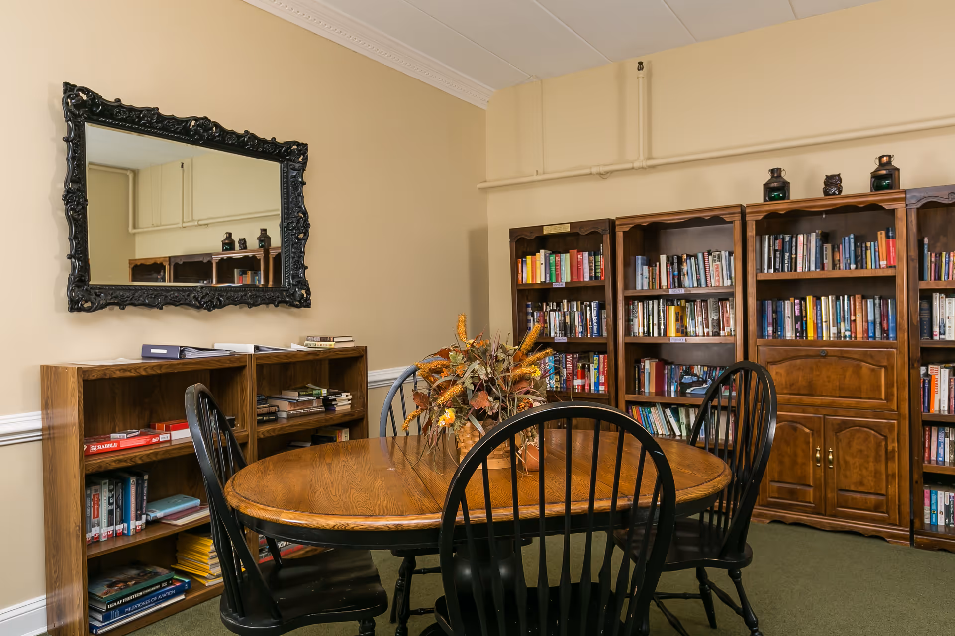 A cozy room with a wooden oval table surrounded by black chairs. Behind the table are several wooden bookshelves filled with books. A decorative mirror with an ornate black frame hangs on the beige wall above a smaller bookshelf. A floral arrangement is placed on the table.