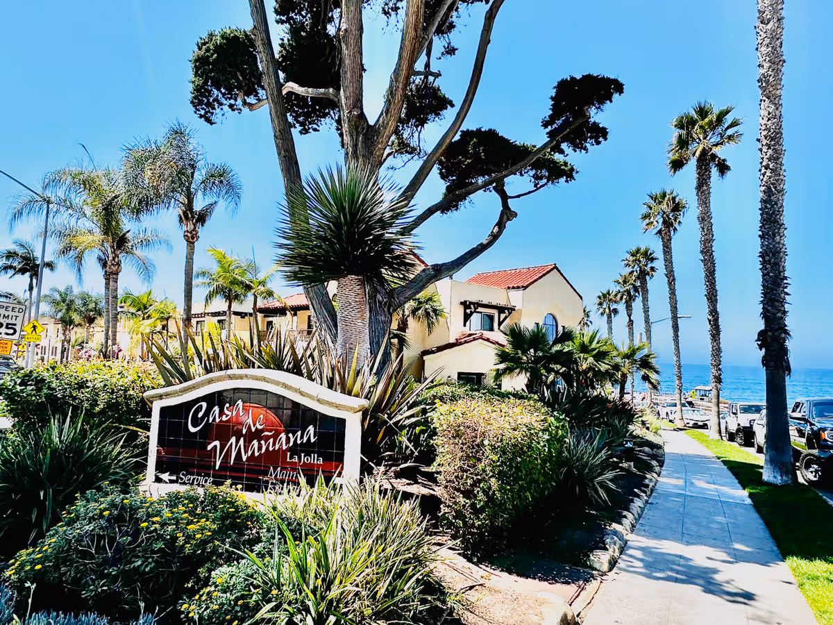 Landscape sign reading "Casa de Mañana" in front of a Spanish-style building with palm trees and the ocean visible in the background.