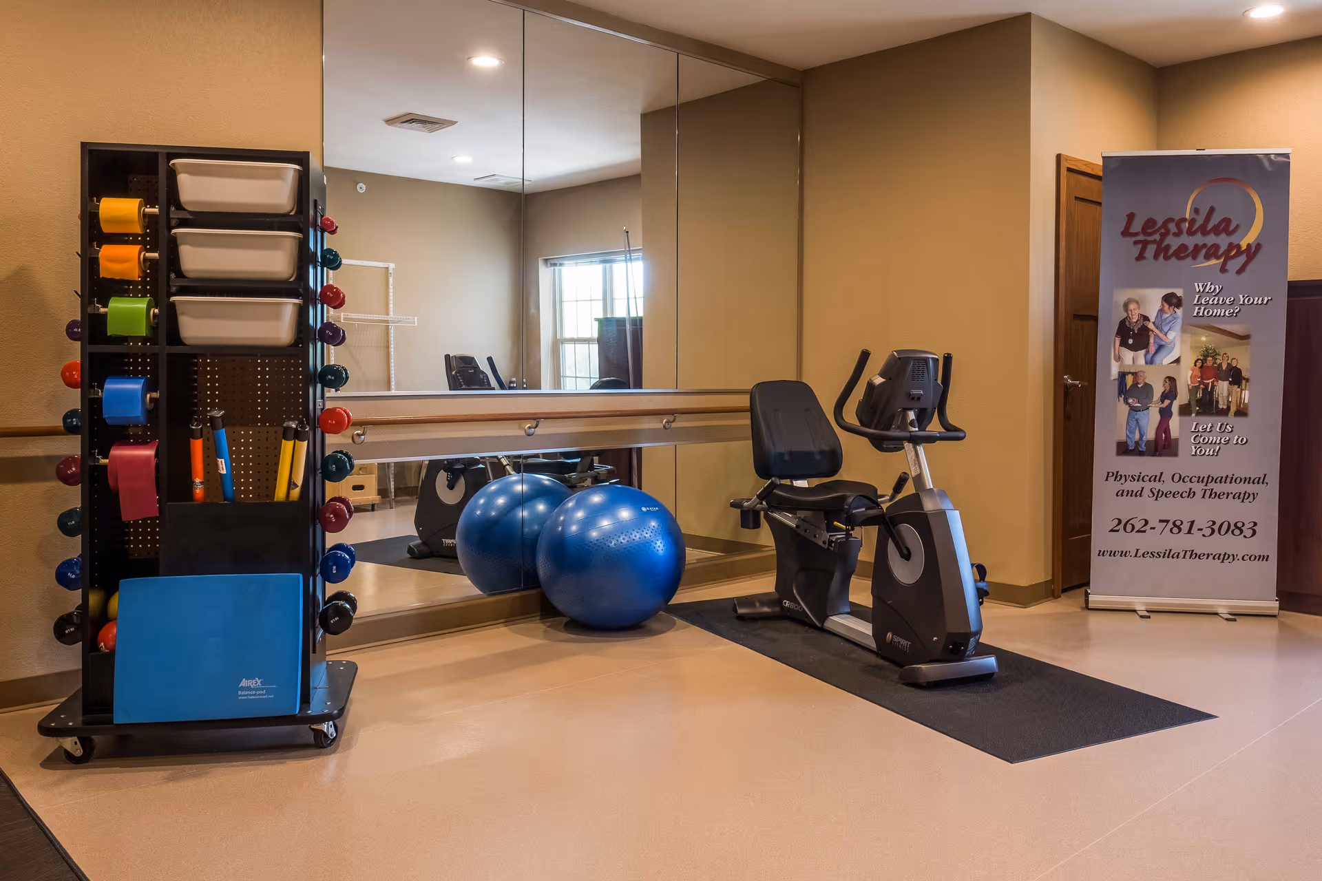 A small exercise room with a stationary recumbent bike on a black mat, two large blue exercise balls, a rack holding colorful resistance bands and small weights, and a large mirror covering one wall. There is a banner in the corner advertising Lessila Therapy for physical, occupational, and speech therapy services.